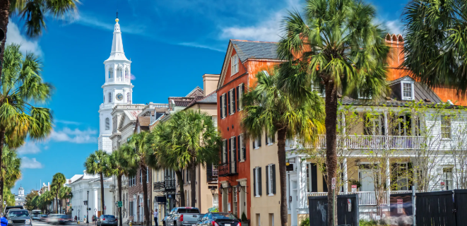 Historic pastel row houses and palmetto trees along a Charleston, South Carolina street, with St. Philip's Church steeple in the background.